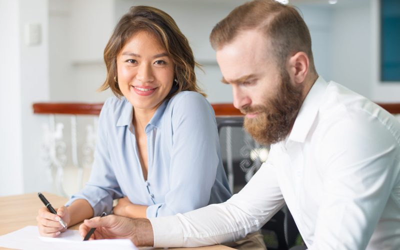 Serious boss checking documents while assistant smiling at camera. Two business colleagues holding pens and working on report. Paperwork concept