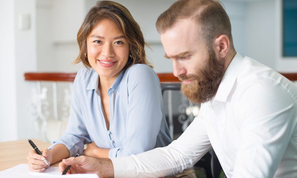 Serious boss checking documents while assistant smiling at camera. Two business colleagues holding pens and working on report. Paperwork concept